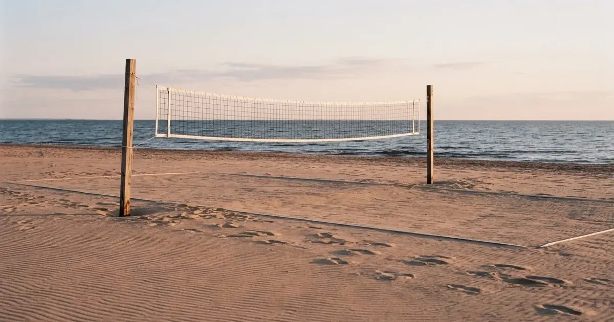 Plażowe boisko do siatkówki o świcie: piaszczysty plac, rozciągnięta siatka i ślady stóp na piasku, w tle morze.